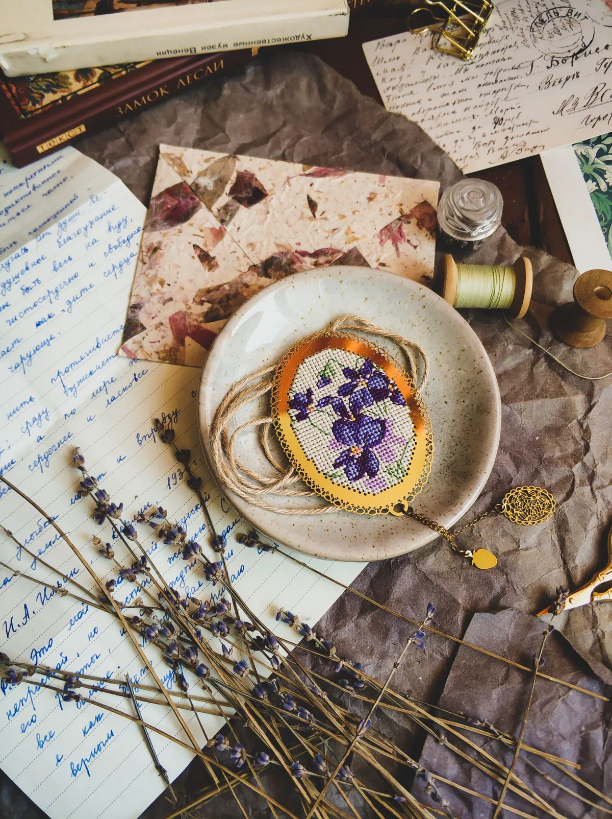 Médaillon brodé de fleurs violettes en point de croix posé sur une assiette, entouré de fils et de papiers vintage