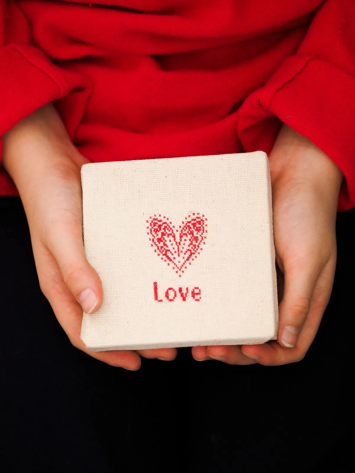 Mains tendant un cadeau brodé point de croix cœur Love — geste d'offrir une broderie faite main personnalisée