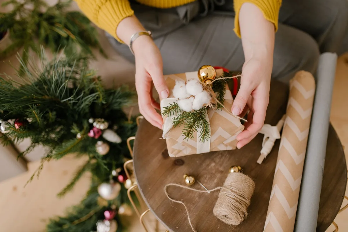 Mains enveloppant un petit cadeau de Noël avec du papier kraft, du coton et une branche de sapin