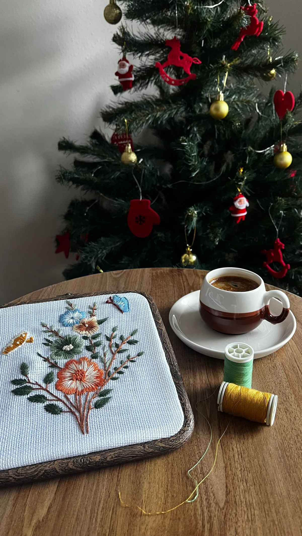 Ouvrage brodé floral posé près d'un sapin de Noël décoré, ambiance cosy avec une tasse de café