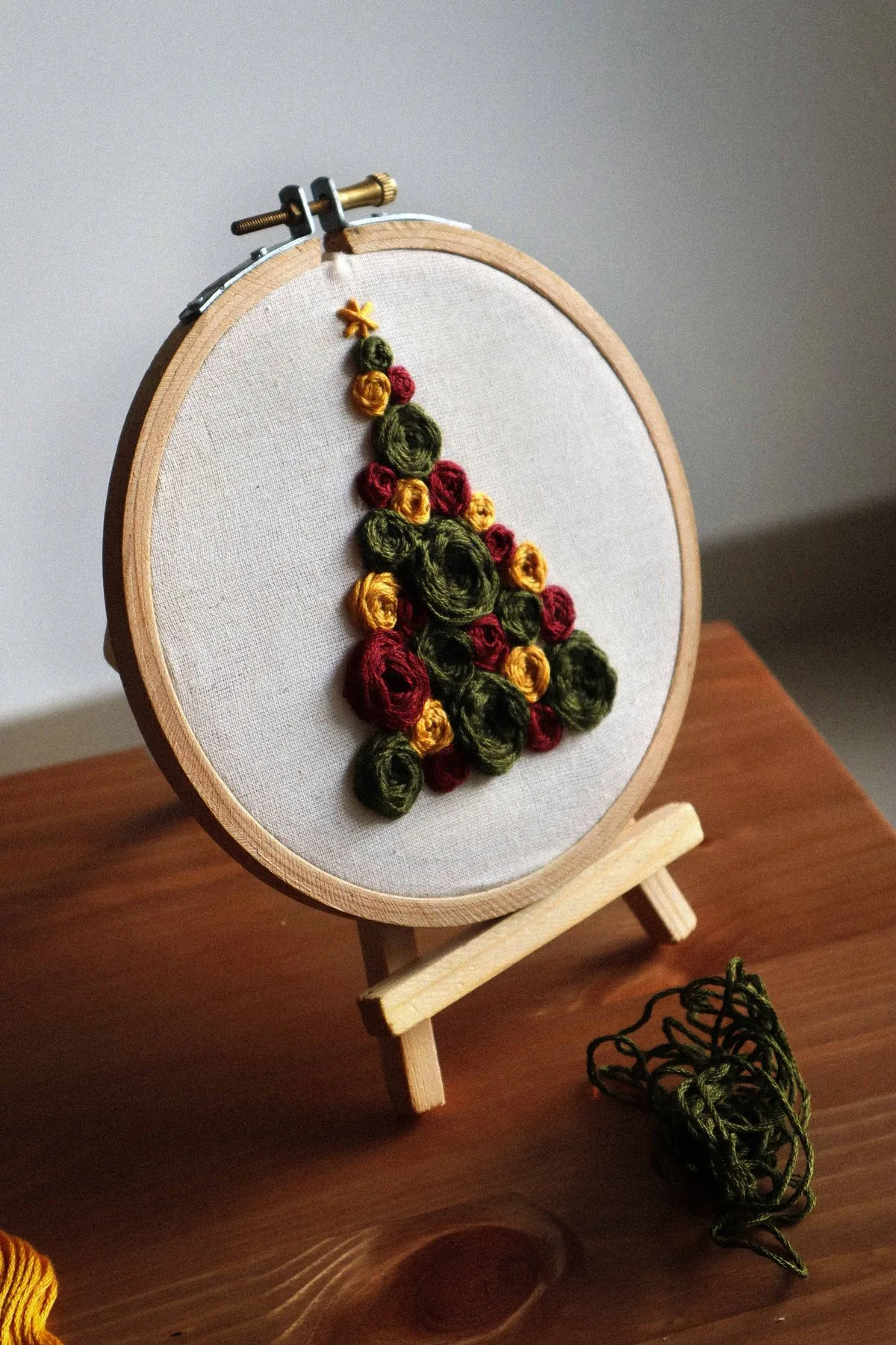 Petit sapin de Noël brodé en fils colorés dans un cerceau miniature, posé sur un chevalet en bois